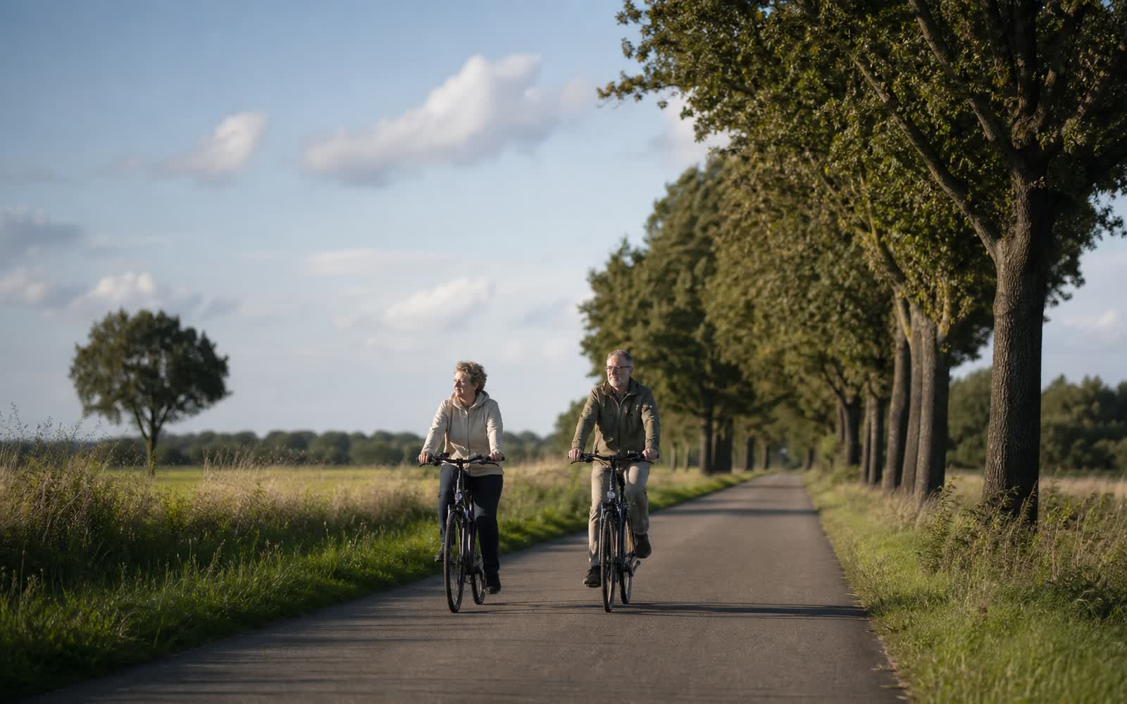 Zwei ältere Erwachsene fahren entspannt mit Fahrrädern über eine Allee am Rand einer flachen norddeutschen Landschaft unter weitem Himmel